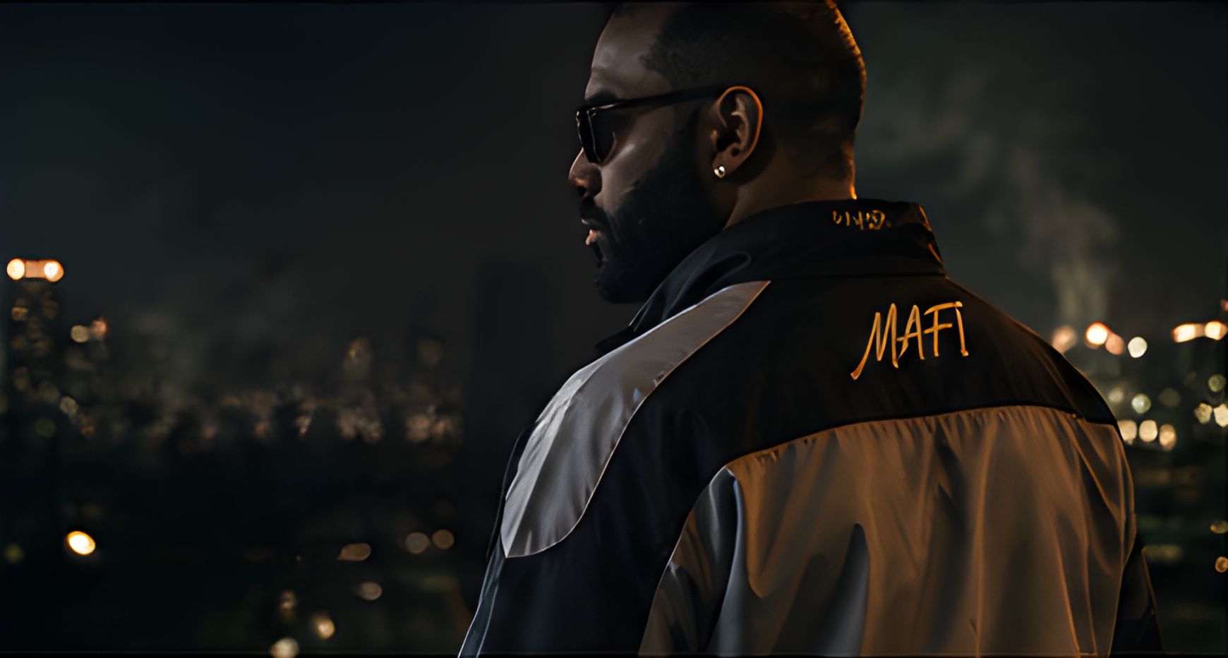 Profile of a man in sunglasses wearing a MAFI windbreaker with the script logo across the back, city lights behind